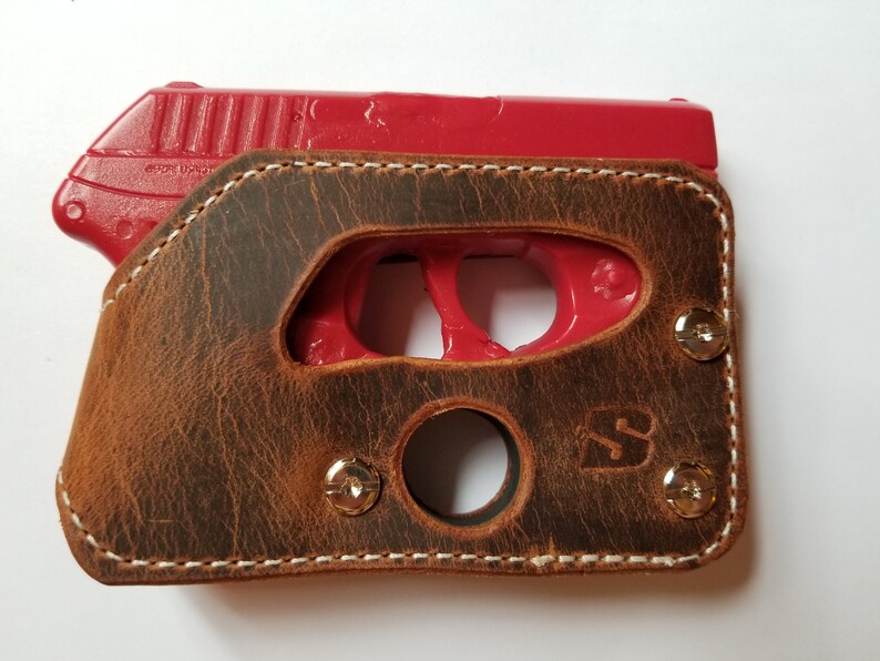 Puede incluir: Una funda de cuero marr&oacute;n con costuras blancas, que sostiene una pistola de entrenamiento roja. La funda tiene un acabado envejecido, remaches met&aacute;licos y un recorte para el gatillo. El cuero est&aacute; estampado con un logotipo.