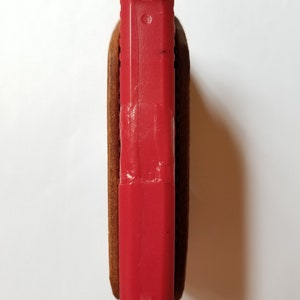 Puede incluir: Una pistola de juguete roja est&aacute; parcialmente encerrada en una funda de cuero marr&oacute;n. La parte superior de la pistola es visible, mostrando las miras y una peque&ntilde;a marca. La funda parece estar hecha de un material de cuero texturizado.