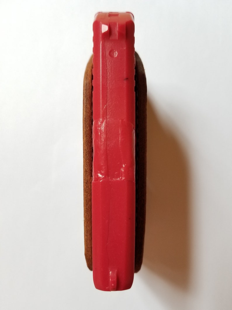 Puede incluir: Una pistola de juguete roja est&aacute; parcialmente encerrada en una funda de cuero marr&oacute;n. La parte superior de la pistola es visible, mostrando las miras y una peque&ntilde;a marca. La funda parece estar hecha de un material de cuero texturizado.