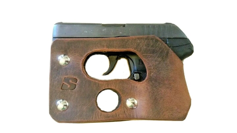 Może przedstawiać: Brązowa sk&oacute;rzana kabura z wycięciem na mały pistolet. Kabura ma metalowe zapięcie na zatrzask i metalowe logo z przodu.