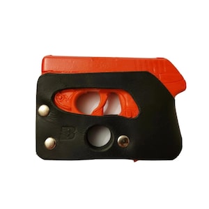Puede incluir: Una funda de cuero negra y naranja para una pistola peque&ntilde;a. La funda es negra con remaches plateados y una abertura circular. El arma de fuego es naranja y visible a trav&eacute;s de las aberturas de la funda.