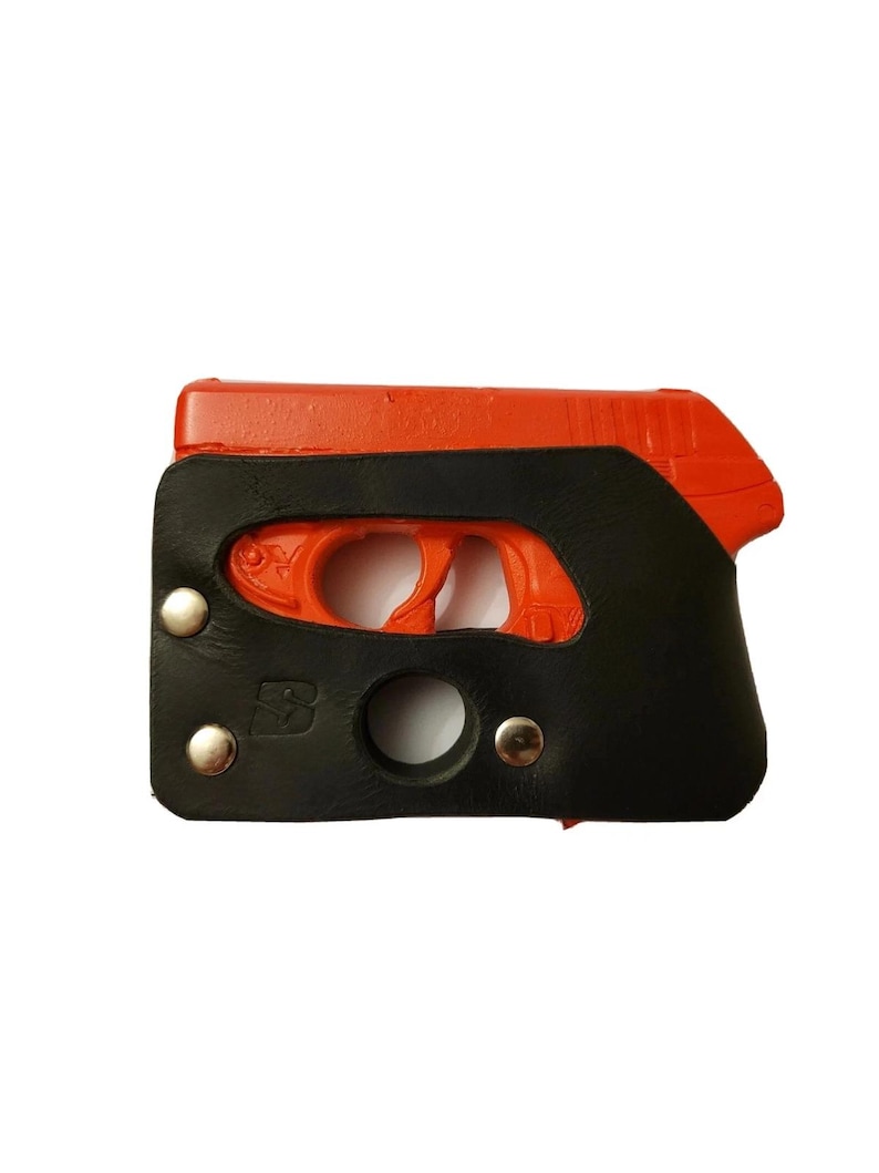 Puede incluir: Una funda de cuero negra y naranja para una pistola peque&ntilde;a. La funda es negra con remaches plateados y una abertura circular. El arma de fuego es naranja y visible a trav&eacute;s de las aberturas de la funda.