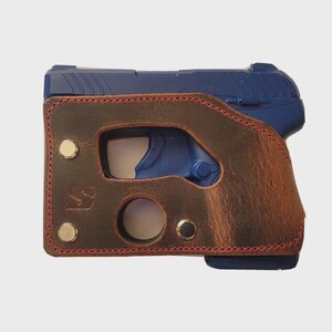 Puede incluir: Una pistola azul est&aacute; parcialmente encerrada en una funda de cuero marr&oacute;n. La funda tiene costuras rojas alrededor de los bordes y est&aacute; asegurada con remaches met&aacute;licos. La funda tiene recortes para el gatillo y el liberador del cargador.