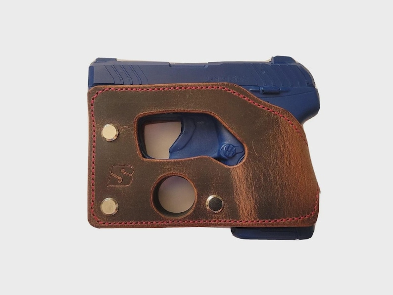 Puede incluir: Una pistola azul est&aacute; parcialmente encerrada en una funda de cuero marr&oacute;n. La funda tiene costuras rojas alrededor de los bordes y est&aacute; asegurada con remaches met&aacute;licos. La funda tiene recortes para el gatillo y el liberador del cargador.