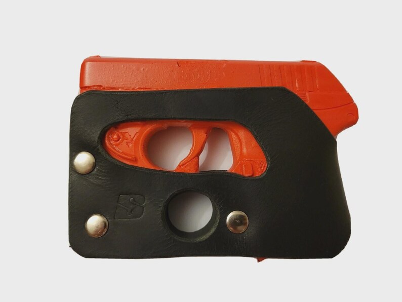 Puede incluir: Una pistola naranja est&aacute; parcialmente encerrada en una funda de cuero negro. La funda est&aacute; asegurada con tres remaches plateados y presenta una abertura circular y un logotipo "S" estilizado. El gatillo y la empu&ntilde;adura de la pistola son visibles.