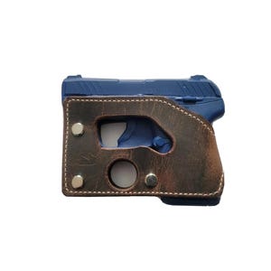 Puede incluir: Una pistola azul est&aacute; dentro de una funda de cuero marr&oacute;n. La funda tiene un borde cosido y est&aacute; asegurada con remaches plateados. El arma es visible a trav&eacute;s de cortes en la funda.