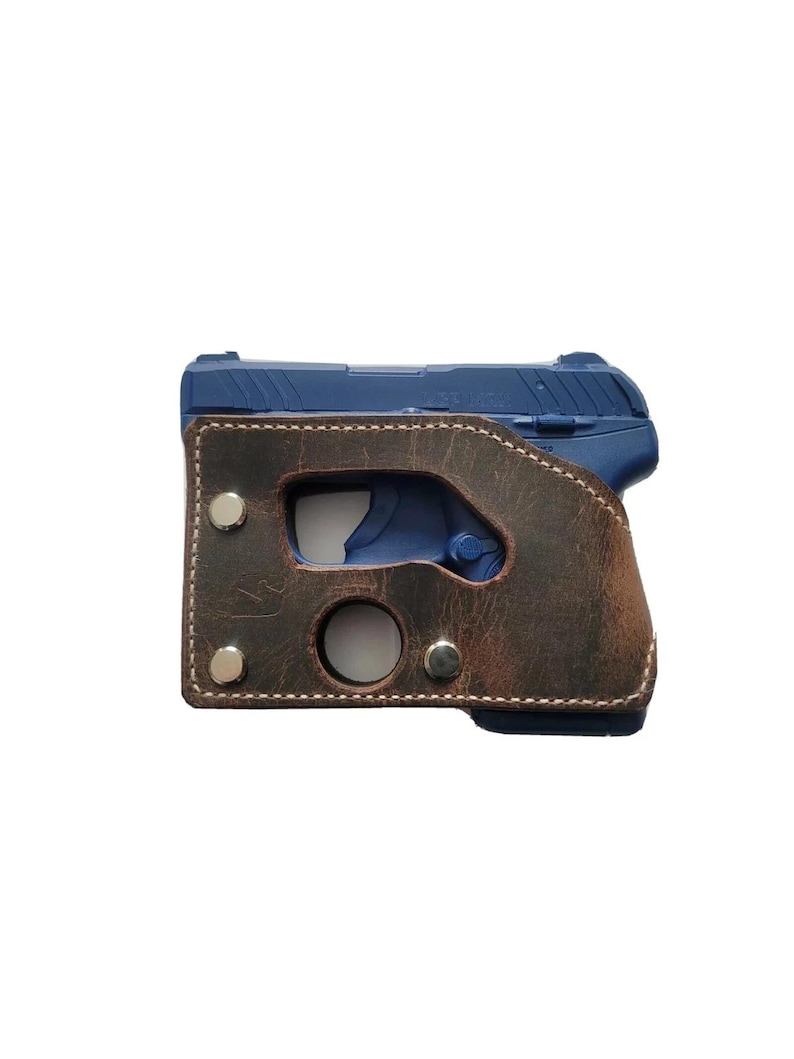 Puede incluir: Una pistola azul est&aacute; dentro de una funda de cuero marr&oacute;n. La funda tiene un borde cosido y est&aacute; asegurada con remaches plateados. El arma es visible a trav&eacute;s de cortes en la funda.