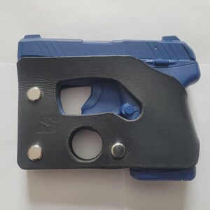 Puede incluir: Una pistola azul en una funda de cuero negro. La funda tiene un recorte para el gatillo y una abertura circular en la parte inferior. Remaches plateados aseguran la funda. El arma es parcialmente visible.