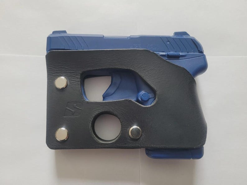 Puede incluir: Una pistola azul en una funda de cuero negro. La funda tiene un recorte para el gatillo y una abertura circular en la parte inferior. Remaches plateados aseguran la funda. El arma es parcialmente visible.
