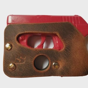 Puede incluir: Una funda de cuero marr&oacute;n con remaches de lat&oacute;n encierra una pistola roja. La funda tiene un acabado envejecido y una abertura circular. El dise&ntilde;o incluye un peque&ntilde;o logotipo y est&aacute; asegurado con tres remaches de lat&oacute;n.