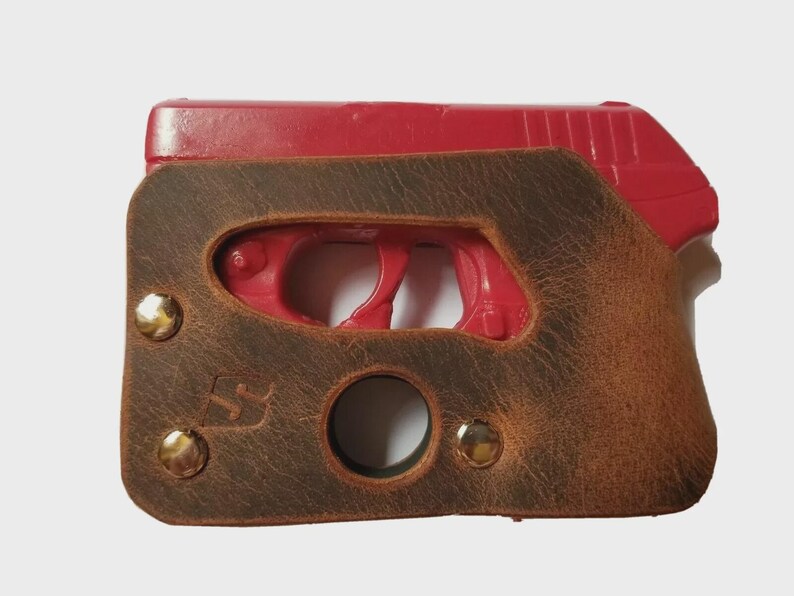Puede incluir: Una funda de cuero marr&oacute;n con remaches de lat&oacute;n encierra una pistola roja. La funda tiene un acabado envejecido y una abertura circular. El dise&ntilde;o incluye un peque&ntilde;o logotipo y est&aacute; asegurado con tres remaches de lat&oacute;n.