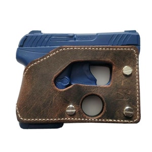Puede incluir: Una pistola azul est&aacute; parcialmente encerrada en una funda de cuero marr&oacute;n. La funda tiene un acabado envejecido, costuras blancas y est&aacute; asegurada con remaches plateados. El arma de fuego es visible a trav&eacute;s de las aberturas de la funda.