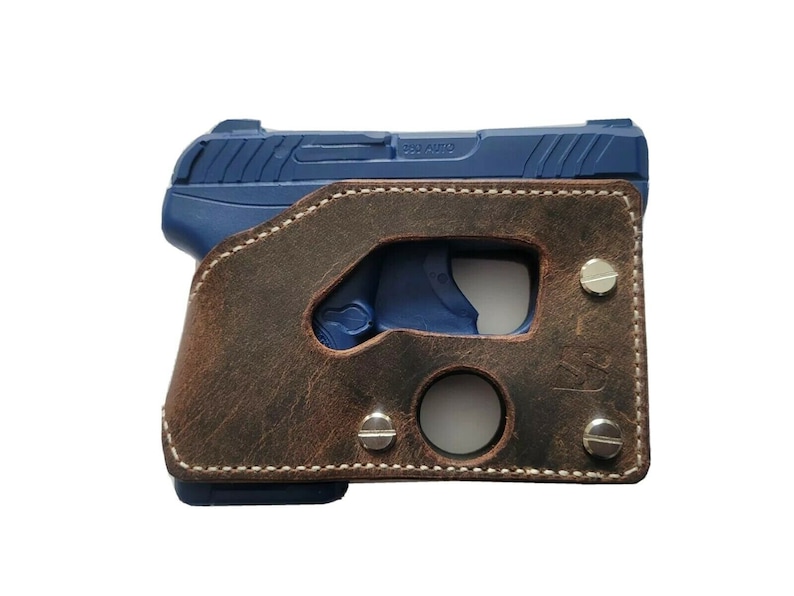 Puede incluir: Una pistola azul est&aacute; parcialmente encerrada en una funda de cuero marr&oacute;n. La funda tiene un acabado envejecido, costuras blancas y est&aacute; asegurada con remaches plateados. El arma de fuego es visible a trav&eacute;s de las aberturas de la funda.