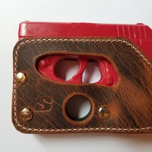 Puede incluir: Una funda de cuero marr&oacute;n con costuras blancas y remaches de lat&oacute;n. La funda est&aacute; dise&ntilde;ada para contener una pistola roja. El cuero tiene un aspecto envejecido y un logotipo visible en la parte delantera.