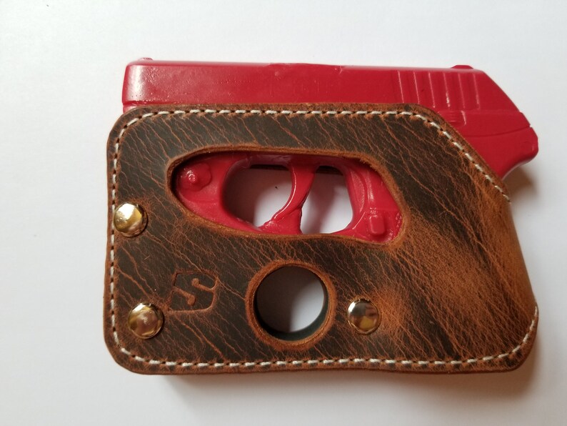 Puede incluir: Una funda de cuero marr&oacute;n con costuras blancas y remaches de lat&oacute;n. La funda est&aacute; dise&ntilde;ada para contener una pistola roja. El cuero tiene un aspecto envejecido y un logotipo visible en la parte delantera.