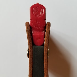 Puede incluir: Una pistola de entrenamiento de pl&aacute;stico rojo se sostiene en una funda de cuero marr&oacute;n con remaches plateados. La funda tiene un interior verde oscuro. La pistola tiene un dise&ntilde;o tallado en la parte superior.
