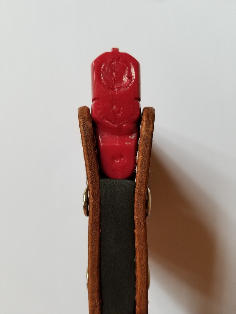 Puede incluir: Una pistola de entrenamiento de pl&aacute;stico rojo se sostiene en una funda de cuero marr&oacute;n con remaches plateados. La funda tiene un interior verde oscuro. La pistola tiene un dise&ntilde;o tallado en la parte superior.