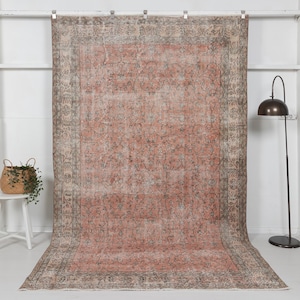 May include: A large, rectangular, vintage rug with a faded, distressed appearance. The rug features a central field of muted coral with an intricate floral pattern. A border of grey and beige frames the design. The rug is displayed against a white wall.