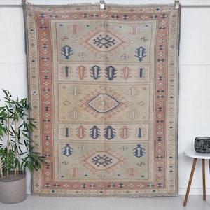 May include: A vintage rug with a beige background and intricate geometric patterns in shades of red, blue, and cream. The rug has a fringed edge and is hanging on a white wall.