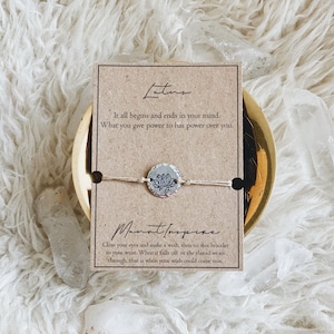 May include: A brown card with the word "Lotus" written in cursive. The card has a quote about the power of the mind. A string bracelet with a silver charm featuring a lotus flower design is attached to the card. The card also has the text "Manifest Inspire" and instructions on how to make a wish with the bracelet.