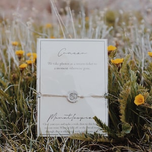 May include: A white card with the text "Camera" and "We take photos as a return ticket to a moment otherwise gone."  The card is sitting on a bed of green grass with yellow flowers. A string bracelet with a silver camera charm is attached to the card.