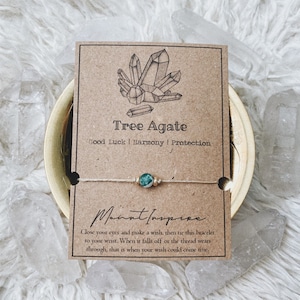 Op de afbeelding: Een groene boom-agaten armband op een wit koord met een kaart waarop staat "Tree Agate Good Luck | Harmony | Protection" en "Sluit je ogen en wens iets, bind dan deze armband om je pols. Wanneer deze eraf valt of de draad versleten is, dan kan je wens uitkomen."