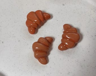 Puede incluir: Tres cruasanes en miniatura de color marrón anaranjado sobre una superficie blanca moteada. Los cruasanes tienen un acabado brillante, con crestas detalladas y una apariencia realista, lo que sugiere una artesanía o decoración con temática de comida.