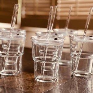 May include: Six clear glass cups with lids and straws. The cups are stacked on top of each other. The cups are on a dark wooden surface.