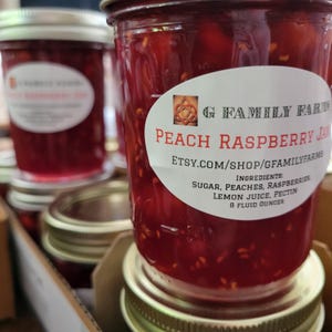 May include: Close-up of several jars of peach raspberry jam. The jam is a vibrant red color and has a chunky texture. Each jar has a white label with the text "Peach Raspberry Jam" and the Etsy shop URL. The ingredients are listed on the label.