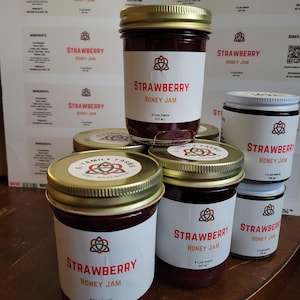 May include: Several glass jars of G. Family Farms Strawberry Honey Jam with gold metal lids. The labels feature a red, green, and black knot logo, the text Strawberry Honey Jam, and volume markings of 8 fluid ounces or 4 fluid ounces.