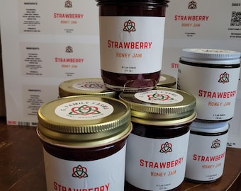 May include: Several glass jars of G. Family Farms Strawberry Honey Jam with gold metal lids. The labels feature a red, green, and black knot logo, the text Strawberry Honey Jam, and volume markings of 8 fluid ounces or 4 fluid ounces.