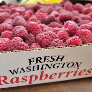 May include: A cardboard box filled with numerous fresh red raspberries. The side of the white box features black text that reads FRESH WASHINGTON and red text that reads Raspberries, indicating the contents of the container.
