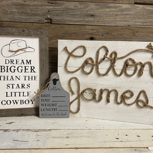 May include: A rustic-themed nursery decor set. It includes a framed sign with a cowboy hat graphic and the text "Dream Bigger Than The Stars Little Cowboy." There's also a wooden sign with the name "Colton James" in rope, and a gray tag.