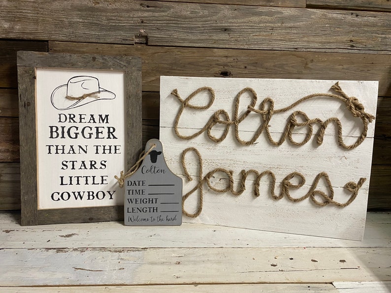 May include: A rustic-themed nursery decor set. It includes a framed sign with a cowboy hat graphic and the text "Dream Bigger Than The Stars Little Cowboy." There's also a wooden sign with the name "Colton James" in rope, and a gray tag.