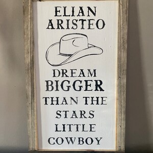 May include: A rectangular wooden sign with a white painted background and a distressed gray and brown frame. The sign features black text and a cowboy hat illustration. The text reads "ELIAN ARISTEO DREAM BIGGER THAN THE STARS LITTLE COWBOY."