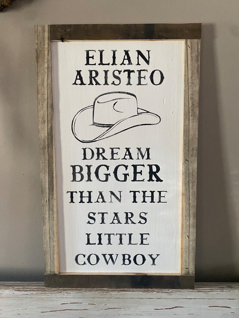 May include: A rectangular wooden sign with a white painted background and a distressed gray and brown frame. The sign features black text and a cowboy hat illustration. The text reads "ELIAN ARISTEO DREAM BIGGER THAN THE STARS LITTLE COWBOY."