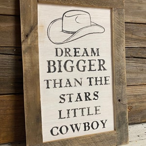 May include: Wooden framed sign with the text "DREAM BIGGER THAN THE STARS LITTLE COWBOY" and a cowboy hat illustration. The sign has a white background and a rustic, weathered wood frame, perfect for a western-themed decor.