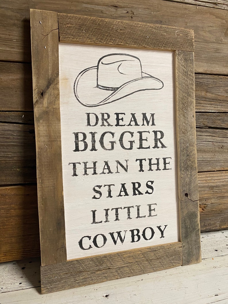 May include: Wooden framed sign with the text "DREAM BIGGER THAN THE STARS LITTLE COWBOY" and a cowboy hat illustration. The sign has a white background and a rustic, weathered wood frame, perfect for a western-themed decor.
