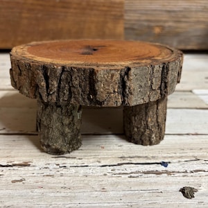May include: A small wooden pedestal with a round top and three legs. The top is made of a natural wood slice with bark still attached. The legs are also made of natural wood with bark still attached.