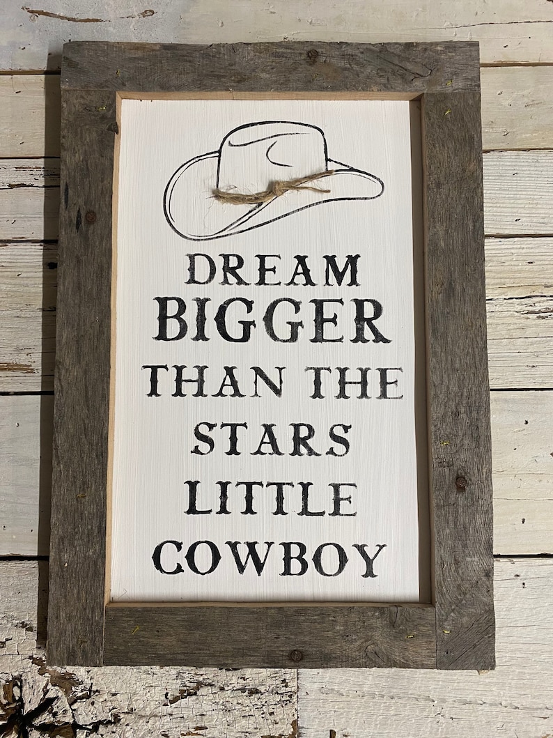 May include: A rectangular wooden sign with a white background and a gray wooden frame. The sign features a black line drawing of a cowboy hat above the words "DREAM BIGGER THAN THE STARS LITTLE COWBOY" in black lettering.
