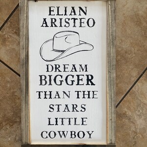May include: A rectangular wooden sign with a white background and a black cowboy hat illustration. The sign reads "ELIAN ARISTEO DREAM BIGGER THAN THE STARS LITTLE COWBOY". The sign is framed with gray wood.