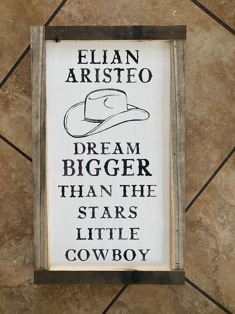 May include: A rectangular wooden sign with a white background and a black cowboy hat illustration. The sign reads "ELIAN ARISTEO DREAM BIGGER THAN THE STARS LITTLE COWBOY". The sign is framed with gray wood.