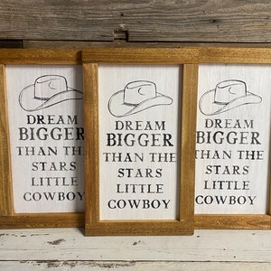 May include: Three framed wooden signs with a white background and a cowboy hat illustration. The signs read "DREAM BIGGER THAN THE STARS LITTLE COWBOY" in black text. The frames are a light brown color.