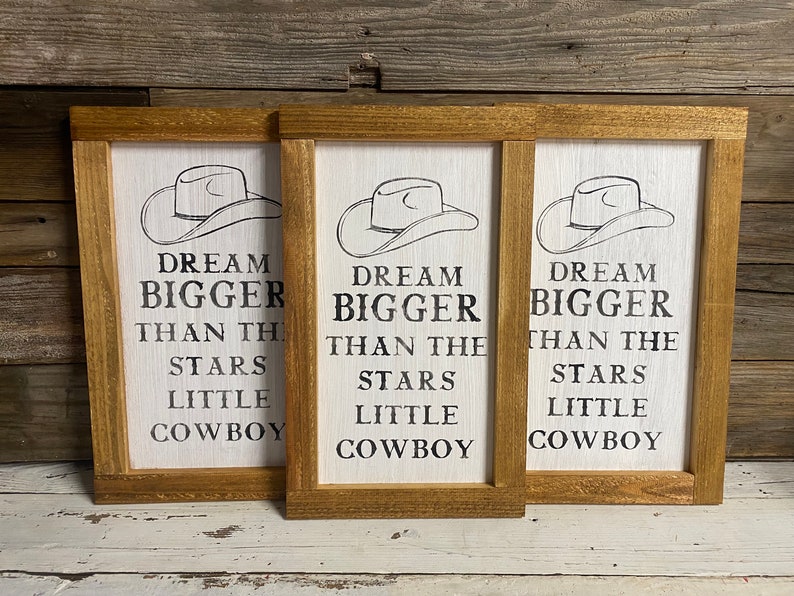 May include: Three framed wooden signs with a white background and a cowboy hat illustration. The signs read "DREAM BIGGER THAN THE STARS LITTLE COWBOY" in black text. The frames are a light brown color.