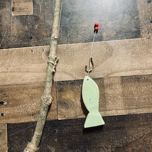 May include: A wooden stick with twine wrapped around the bottom, used as a fishing rod. A thin string with red beads and a hook is attached to the top of the stick. A green wooden fish is hanging from the hook.