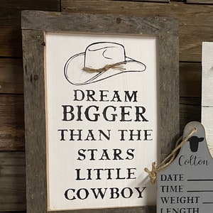 May include: A framed wooden sign with the text "DREAM BIGGER THAN THE STARS LITTLE COWBOY" in black lettering. The sign features a cowboy hat illustration and a rustic wooden frame. A gray tag with the name "Colton" and other details hangs nearby.