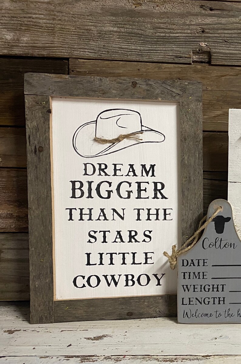 May include: A framed wooden sign with the text "DREAM BIGGER THAN THE STARS LITTLE COWBOY" in black lettering. The sign features a cowboy hat illustration and a rustic wooden frame. A gray tag with the name "Colton" and other details hangs nearby.