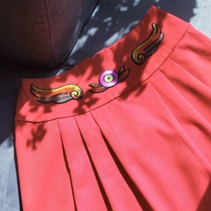 Peut inclure: Jupe corail plissée avec un motif brodé à la taille. Le motif comprend deux ailes dorées et orange et un élément central circulaire avec des accents roses et violets. La jupe est en tissu lisse.