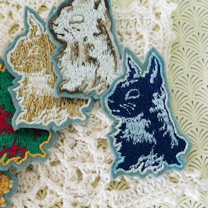 May include: A collection of embroidered bunny patches in shades of gold, green, brown, and navy. Each patch depicts a detailed rabbit design with a light blue border. The patches are displayed on a white, lacy surface.