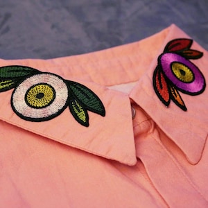 Peut inclure: Gros plan sur un col de chemise rose clair orné de broderies florales. La broderie présente un motif en forme d'œil blanc, jaune et vert d'un côté et un motif violet, orange et jaune de l'autre. La chemise est boutonnée.